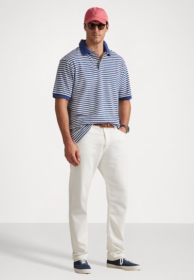 Vīrietis ar zilu un baltu svītrojumu polo krekls, baltām biksēm, rozā cepuri, saulesbrillēm, brūnu pītu jostu, pulkstenis un tumši zilas sporta kurpes, stāv ar rokām kabatās.