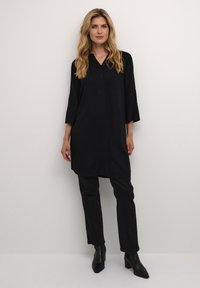 Tunique noire avec un décolleté en V et des manches trois-quarts, associée à un pantalon noir et des bottines à talons, le tout sur un fond neutre.