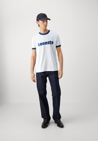 Camiseta blanca con cuello azul y logo de "Lacoste", combinada con jeans de mezclilla oscura con costuras en azul claro y zapatos negros.
