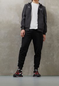 Sudadera negra con cremallera y detalles en blanco, combinada con pantalones de chándal negros ajustados. Camiseta blanca debajo y zapatillas negras y rojas con detalles estampados.