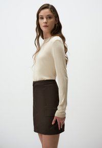 Beige sweater met lange mouwen gecombineerd met een donkerbruine mini-jurk met een voorkantzak, een gladde textuur en een aansluitende pasvorm, weergegeven in profiel.