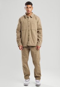 Giacca khaki con due tasche sul petto, chiusura con bottoni e colletto, abbinata a pantaloni coordinati. Entrambi i capi presentano una consistenza liscia e durevole.