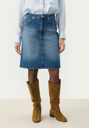 Personne portant une jupe en jean bleue jusqu'aux genoux, une chemise blanche boutonnée, des collants noirs et des bottes en daim marron jusqu'aux genoux, debout sur un sol uni.