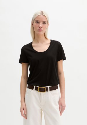Femme aux cheveux blonds lisses portant un t-shirt noir à manches courtes, un pantalon blanc et une ceinture marron à boucle dorée, debout devant un fond blanc.