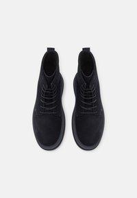 Botas de tobillo de ante negro con punta redondeada, cierre de cordones en la parte delantera y cuello acolchado. Presenta una suela texturizada y un detalle de marca mínimo.
