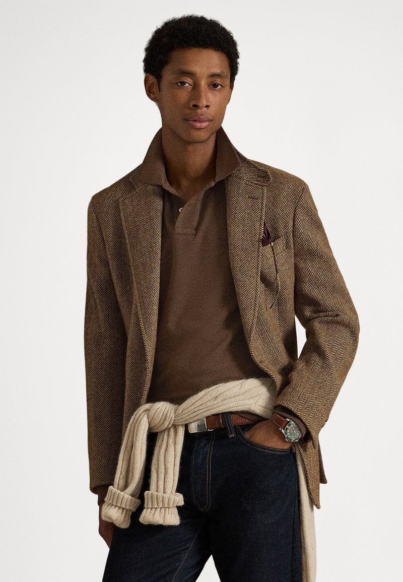 Braunes Fischgrätenmuster-Blazer mit gekerbtem Revers, getragen über einem braunen Poloshirt. Cremefarbener gestrickter Pullover, in der Taille gebunden, kombiniert mit dunklen Jeans.