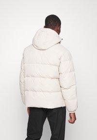 Hombre que lleva una chaqueta de plumas beige clara con capucha, visto desde atrás, combinado con pantalones negros sobre un fondo sencillo.