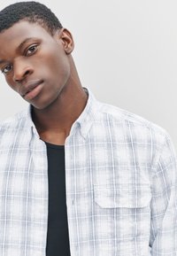 Camicia a quadri bianca e blu con bottoni e colletto, caratterizzata da una tasca sul petto e una parte superiore con sfondo nero semplice. Tessuto dalla texture liscia.