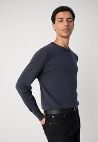 Tengerészkék kötött pulóver kerek nyakkal, slim fit szabással és hosszú ujjal; fekete farmerral és fekete övvel párosítva. Simaszerű textúra, minimalista kialakítás.