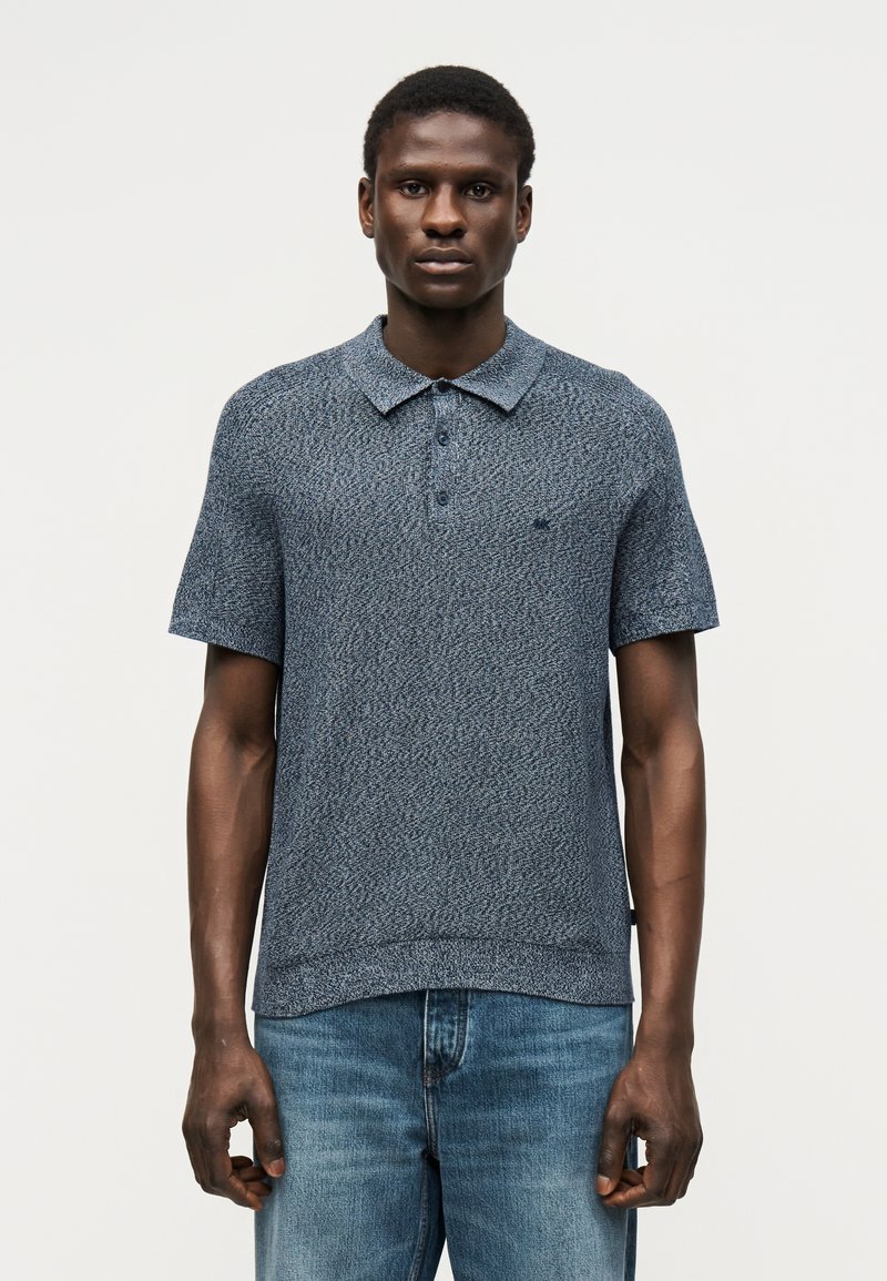 Homme portant un polo bleu texturé à manches courtes et un jean bleu, debout devant un fond clair uni.