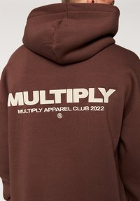 Felpa con cappuccio marrone e una texture morbida, con grande testo bianco "MULTIPLY" e "MULTIPLY APPAREL CLUB 2022" sul retro.