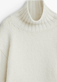 Pull-over blanc à col roulé fabriqué en maille texturée, avec un col à côtes haut et un design à épaules tombantes.