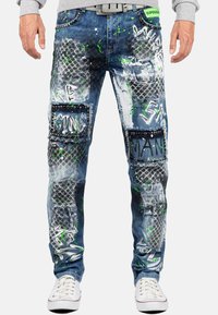 Pantalons en denim pour hommes avec un design éclaboussé de peinture bleue et blanche, des patchs texturés et des accents cloutés, accompagnés de texte en style graffiti vert et noir.
