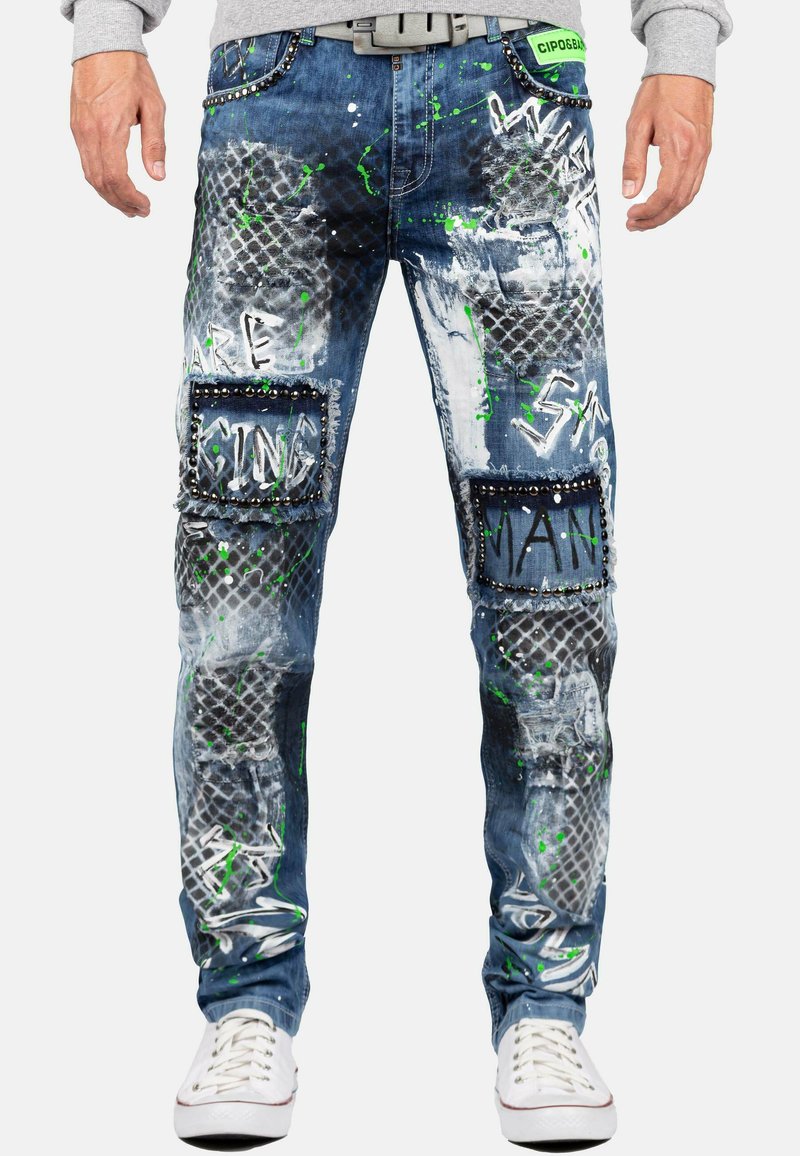 Pantalons en denim pour hommes avec un design éclaboussé de peinture bleue et blanche, des patchs texturés et des accents cloutés, accompagnés de texte en style graffiti vert et noir.