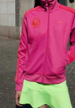 Personne portant une veste zippée rose Adidas Originals avec des bandes orange et une jupe vert vif, debout à l'extérieur sur un pavé en pierre.
