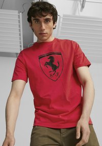 Czerwony bawełniany t-shirt z czarnym emblematem skaczącego konia w obrębie tarczy, towarzyszącym literom "S F". Krótkie rękawy i luźny krój.