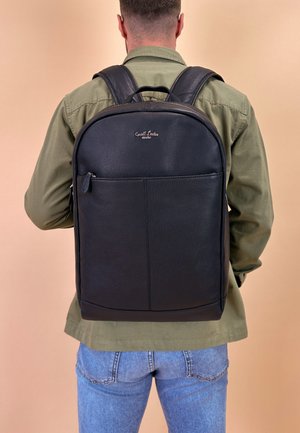 Zaino in pelle nera con superficie liscia, tasca anteriore con zip e spallacci regolabili. Logo visibile sul pannello frontale superiore. Maniglia sulla parte superiore.