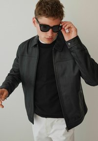Schwarze Lederjacke mit Reißverschluss, Druckknopfkragen und klaren Linien, getragen über einem schwarzen Pullover, kombiniert mit dunklen Sonnenbrillen.