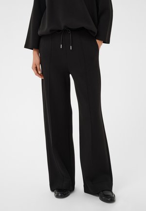 Personne portant un pantalon noir à jambes larges avec des coutures avant, un haut noir et des chaussures noires, debout devant un fond blanc.