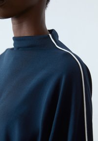 Marineblå høyhalset topp med hvite detaljer. Myk, teksturert stoff faller jevnt, med et minimalistisk design og moderne silhuett.