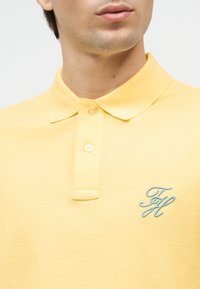 Άνδρας που φοράει ανοιχτό κίτρινο πόλο με δύο κουμπιά και μικρό μπλε κεντημένο λογότυπο "FH" στο στήθος.
