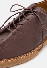 Brown leather shoe featuring a textured surface, round toe, and laces. The sole is light brown with a contrasting stitching detail.