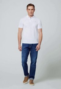 Man som står klädd i en vit polo, blå jeans och bruna sneakers mot en enkel ljusgrå bakgrund.