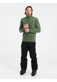 Pullover verde in fleece con colletto a mezza zip, maniche lunghe e logo discreto, abbinato a pantaloni da sci neri e scarponi da sci neri.