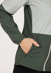Veste légère avec un design en blocs de couleurs gris et vert foncé, dotée d'une poche zippée et de poignets élastiques pour un ajustement parfait.