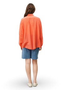 Langärmliges, korallenfarbenes Shirt mit lockerer Passform, aus leichtem Stoff. Kombiniert mit Jeansshorts, die einen fransigen Saum aufweisen.