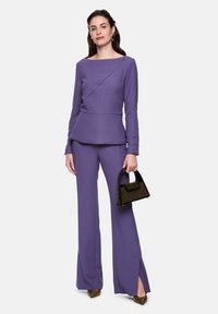 Purple, long-sleeve top with an asymmetric design and peplum waist, paired with matching flared pants. Handholding a small black handbag.
