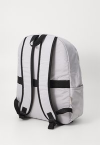 Mochila de tecido cinza com alças pretas, de formato arredondado e um bolso lateral. Apresenta um robusto puxador na parte superior e alças de ombro ajustáveis.