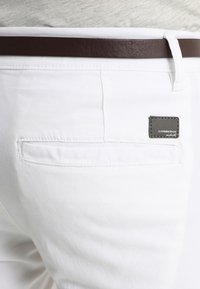 Pantalones de algodón blanco con un corte ajustado, que presentan un cinturón de cuero marrón y una etiqueta gris en el bolsillo trasero. Textura suave, diseño minimalista.