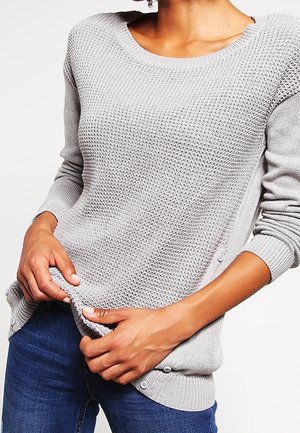 Femme portant un pull en maille texturée gris clair avec des boutons sur les côtés, associé à un jean bleu foncé, tenant l'ourlet du pull avec les deux mains.