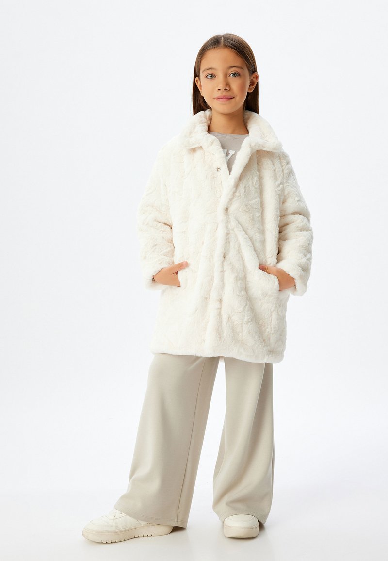 Cappotto in pelliccia sintetica bianca con colletto, maniche lunghe e due tasche laterali. Indossato sopra una camicia grigia chiara e pantaloni a gamba larga beige con sneakers bianche.
