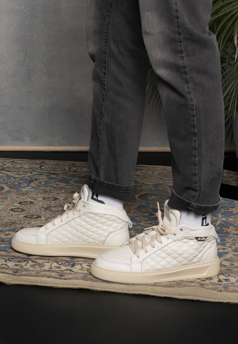 White quilted sneakers with beige accents and light-colored rubber soles, paired with gray jeans and white socks featuring black stripes.