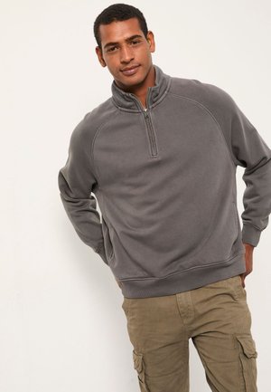 Mann trägt einen grauen Pullover mit Viertelreißverschluss und beige Cargohosen, steht mit den Händen hinter dem Rücken vor einem einfarbigen Hintergrund.