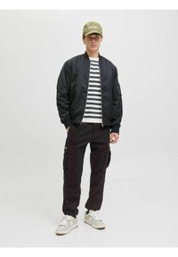 Schwarze Bomberjacke, gestreiftes weißes und marineblaues Hemd, dunkle Cargo-Hosen und eine hellgrüne Kappe. Die Turnschuhe haben graue und weiße Akzente.