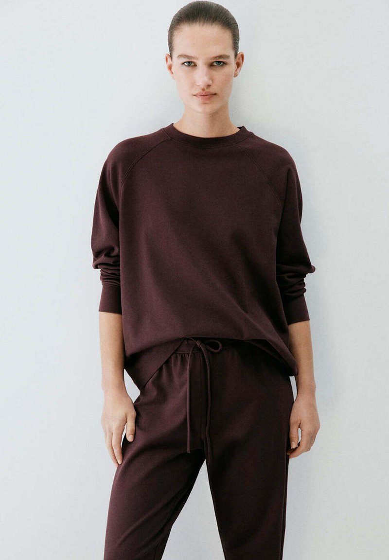 Femme debout devant un fond uni portant un haut ample marron foncé à manches longues et un pantalon à cordon assorti.