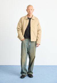 Man standing with one hand in pocket wearing a beige jacket, black shirt, loose faded jeans, and black sneakers on a blue floor.