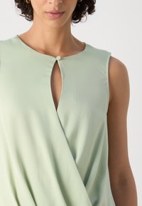 Haut sans manches vert clair en tissu texturé, avec un design portefeuille et un petit bouton décoratif au niveau de l'encolure.