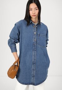 Prevelik denim srajca v modri barvi z gumbnim zapiranjem spredaj, žeparenjem in zavihanimi rokavi. Model drži majhno rjavko torbico.