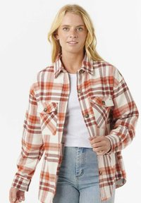 Blonde Frau, die ein weißes Cropped-Top, hochtaillierte hellblaue Jeans und ein übergroßes Hemd mit orange-braunem Karomuster trägt.