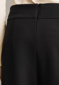 Pantalons noirs taille haute en tissu lisse, avec une ceinture bien structurée et une fermeture éclair dissimulée à l'arrière.