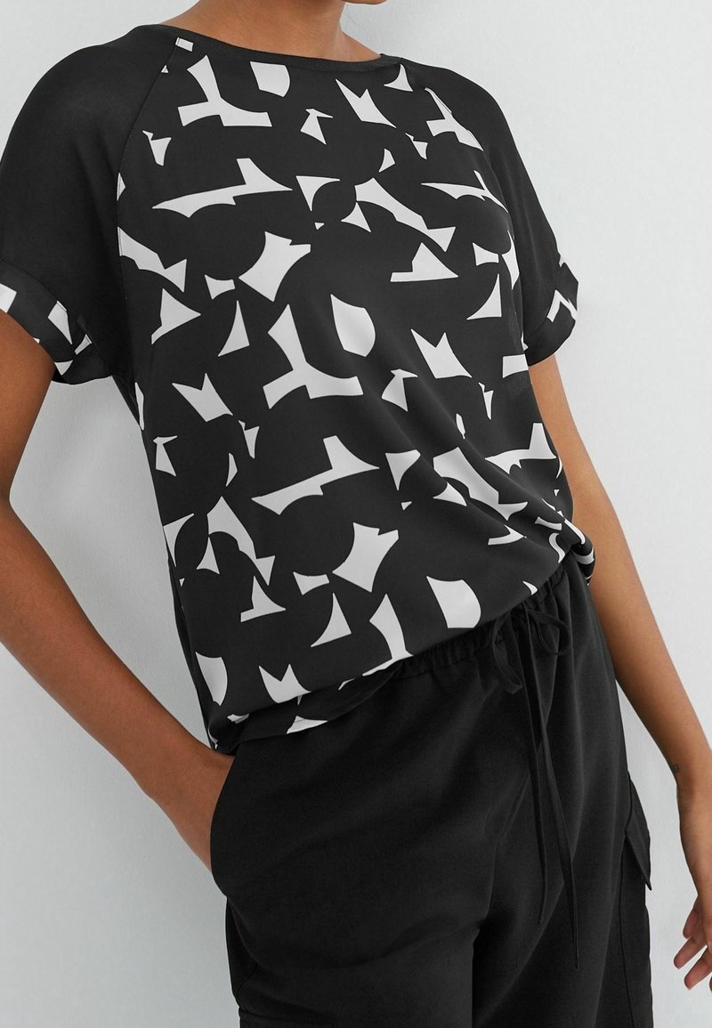 Personne portant un haut noir à manches courtes avec des formes abstraites blanches et un pantalon noir à cordon, debout avec une main dans la poche.