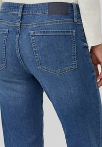 Jeans en denim bleu moyen avec une coupe ajustée, présentant une étiquette de ceinture noire, des poches arrière et des détails de couture subtils.