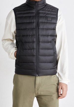 Mężczyzna w czarnej pikowanej kamizelce bez rękawów z zamkiem błyskawicznym i przednimi kieszeniami, noszący białą koszulę z długim rękawem oraz oliwkowe spodnie.