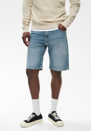 Man met lichtblauwe denim shorts, beige trui, witte sokken en zwarte sneakers, staand met één hand in de zak.