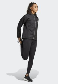 Giacca leggera nera con chiusura a zip e cappuccio, abbinata a leggings neri a vita alta e scarpe da ginnastica nere. Sfondo neutro.