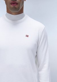Camicia bianca a maniche lunghe con scollatura rotonda, caratterizzata da una piccola bandiera norvegese ricamata sul petto. Tessuto morbido, materiale in cotone.
